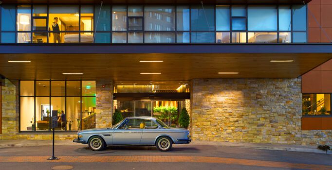 A classic car sits outside of a modern hotel at dusk.