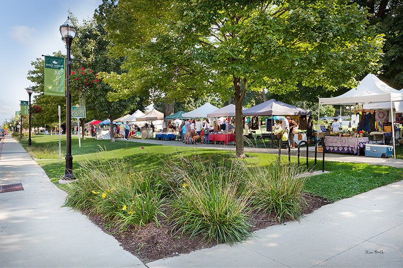 A row of vendor tents line a street at a farmers market on a warm, sunny day.