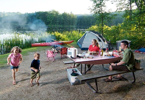 Four people play in a campsite with a tent, kayak, picnic table, and camp fire on a warm day.