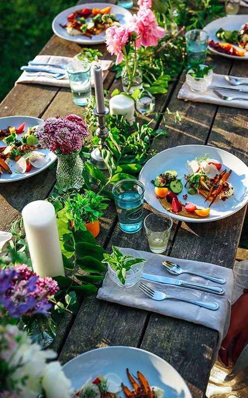 Seen from above, a group of place settings sit on a wooden table outdoors on a warm sunny day.
