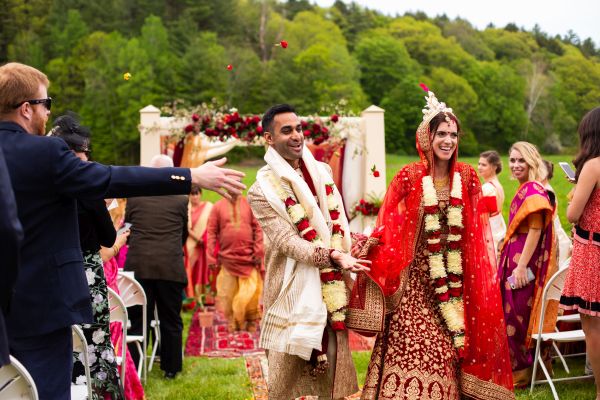 A bride and groom exit their ceremony wearing colorful red and gold clothing. The guests toss flower petals at the couple.