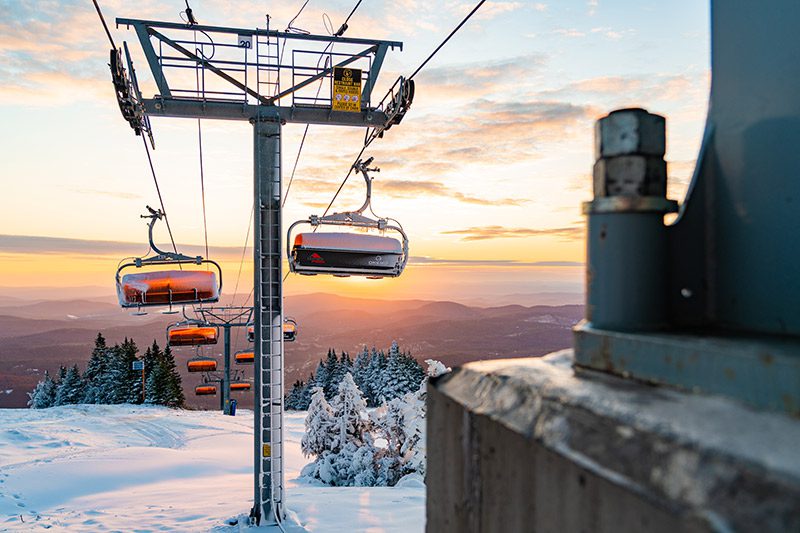 The sun rises behind a char lift at a snowy ski resort.