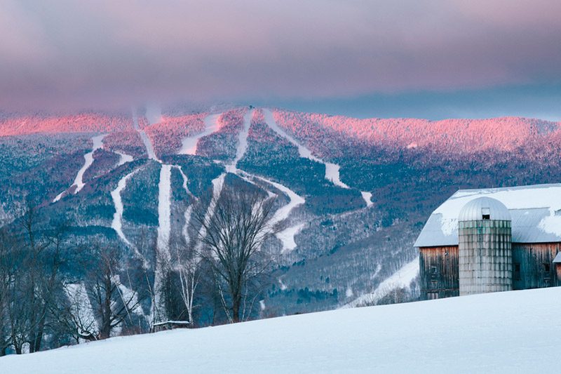A snowy ski resort is seen at sunrise with a barn in the foreground.