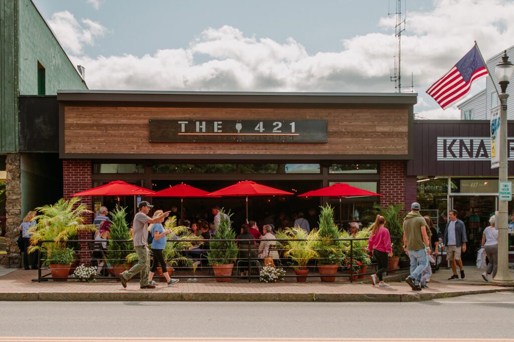 People walk by the front of a restaurant where diners are eating outside under red umbrellas.