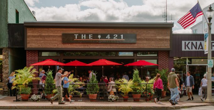 People walk by the front of a restaurant where diners are eating outside under red umbrellas.