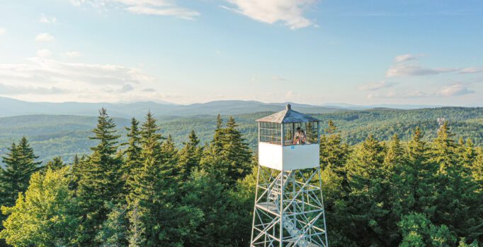Two people stand in a fire tower that extends above the tree line at the top of a mountain on a warm day.