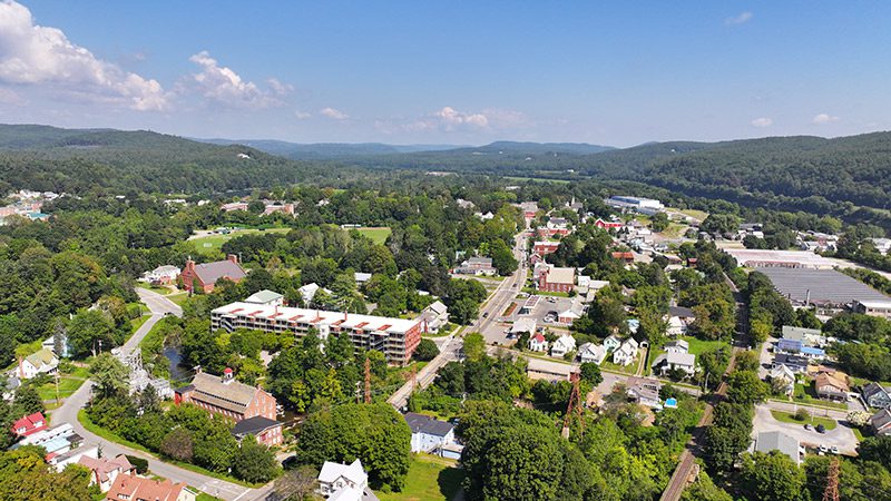 Seen from above, a rural town is nestled between trees and mountains on a warm and sunny day.