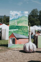 A painting of a red barn is presented outdoors on a sunny day next to a large sculpture of a clove of garlic.