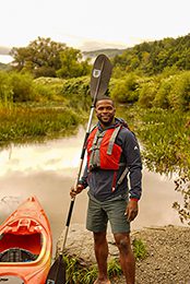 A person in an orange vest stands next to an orange canoe by a river. They are holding a paddle upright and smiling.