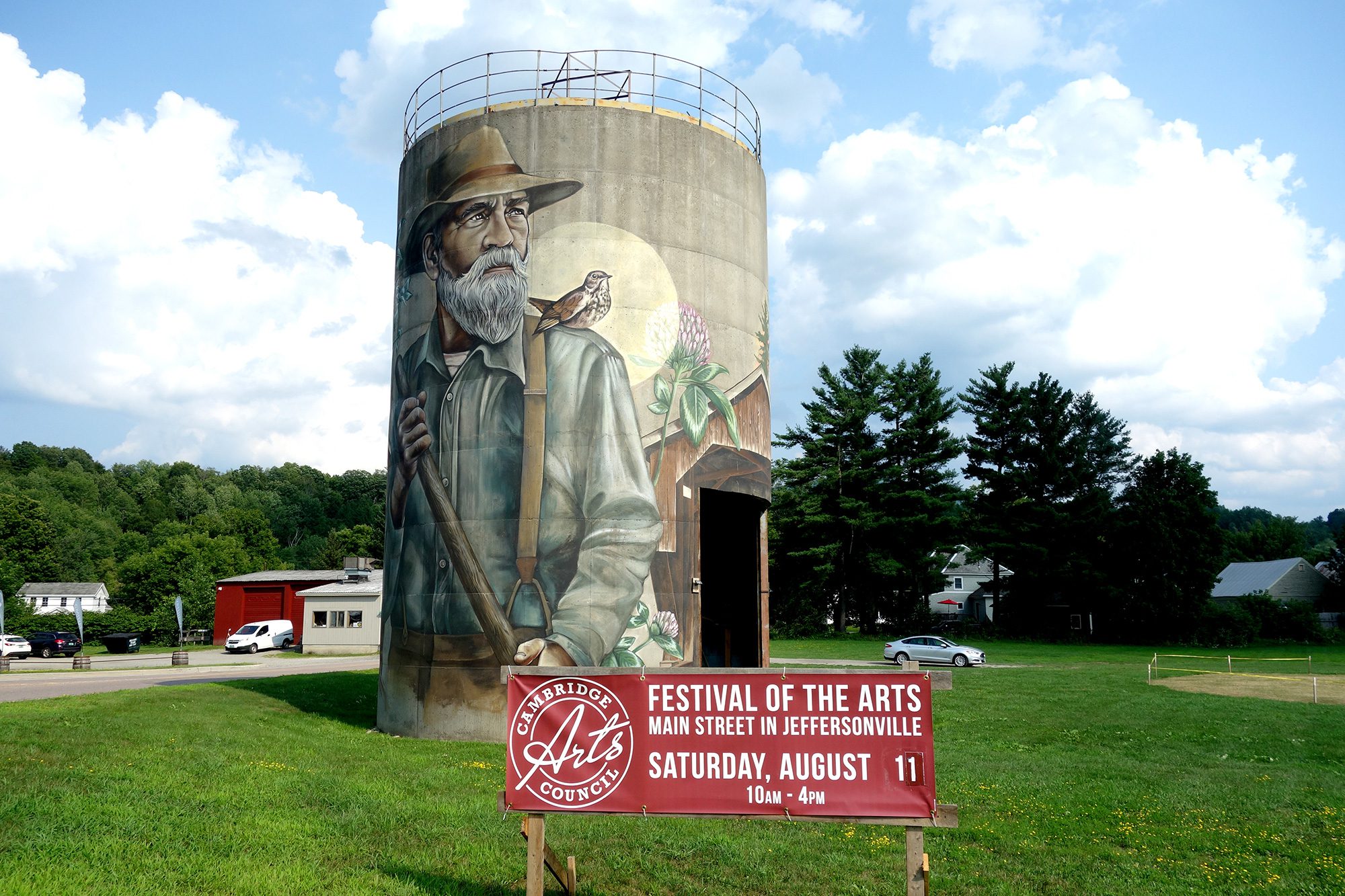 A mural depicting an old man wearing a wide brimmed hat is painted on the side of a silo in a field. 