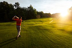 Seen from behind, a person swings a golf club at a golf course while the sun reflects brightly in the distance.