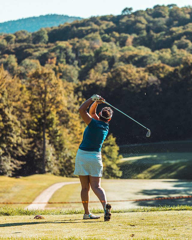 Seen from behind, a person swings a golf club while warm sunlight shines on the golf course.