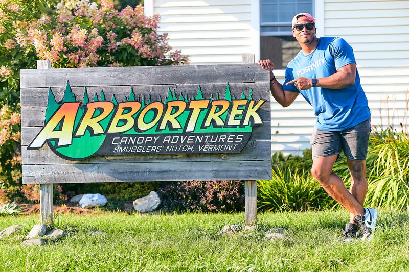 A colorful sign reading ArborTrek with a person leaning against it and smiling.