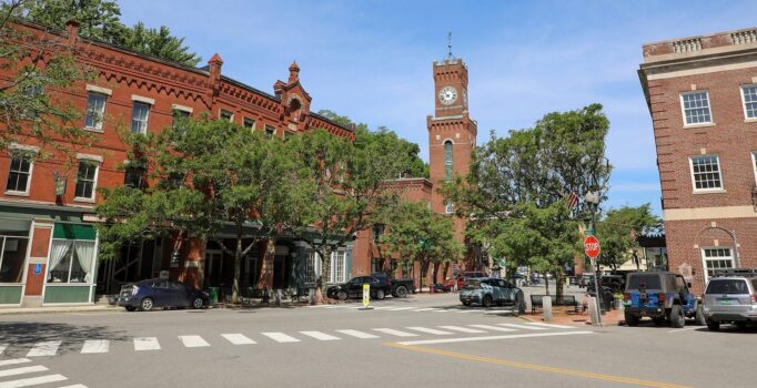 Brick buildings with store fronts, and a tall brick clock, line a paved street on a warm day.