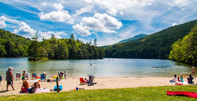 People of all ages lounge and play on a sandy beach at a lake with a forest on the other side.