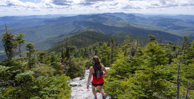 A person seen from the summit climbs down a mountain peak with other mountains visible in the background on a summer day. 