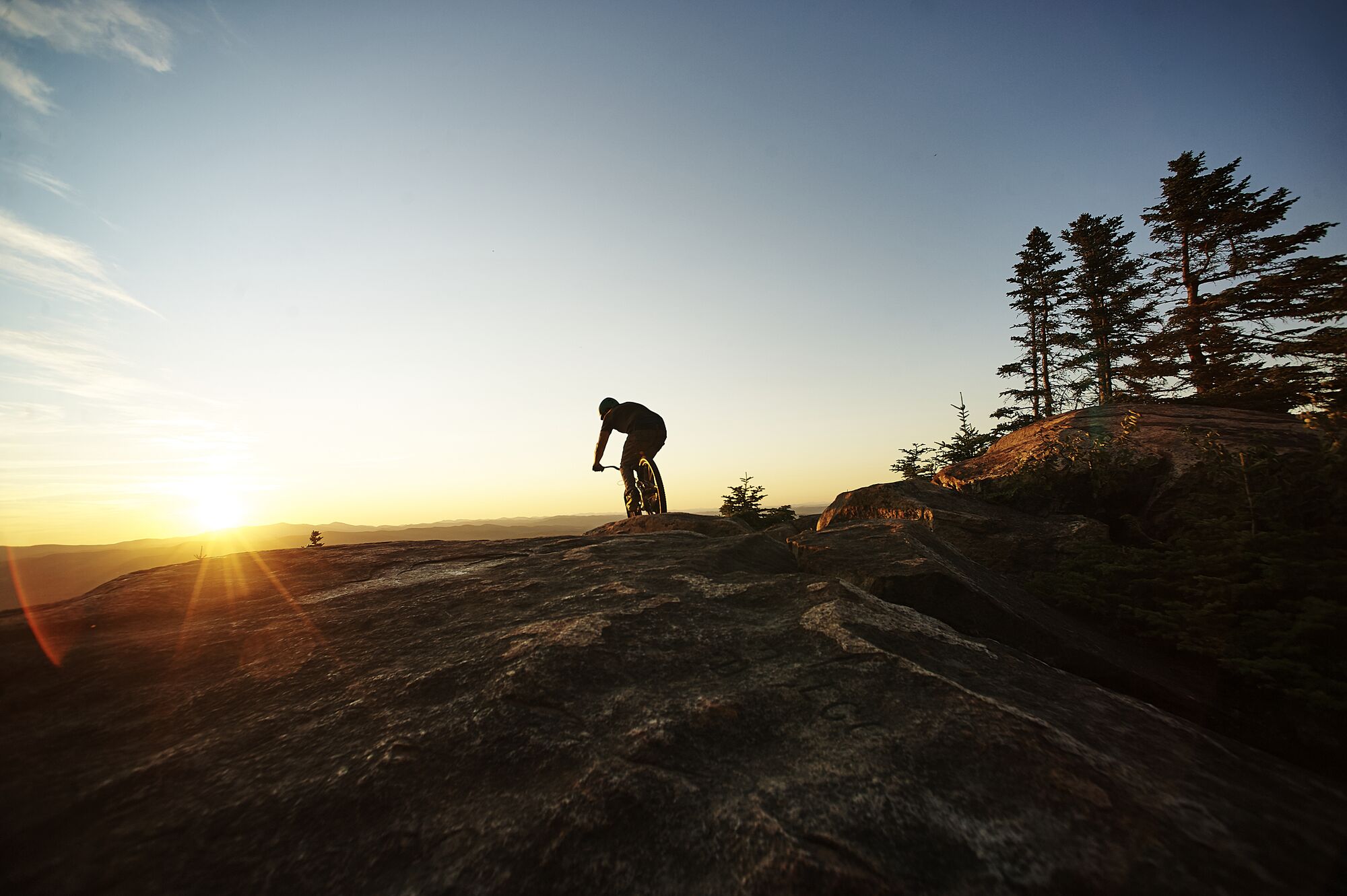 A person rides a bike on the rocky peak of a mountain with the sun setting in the background.