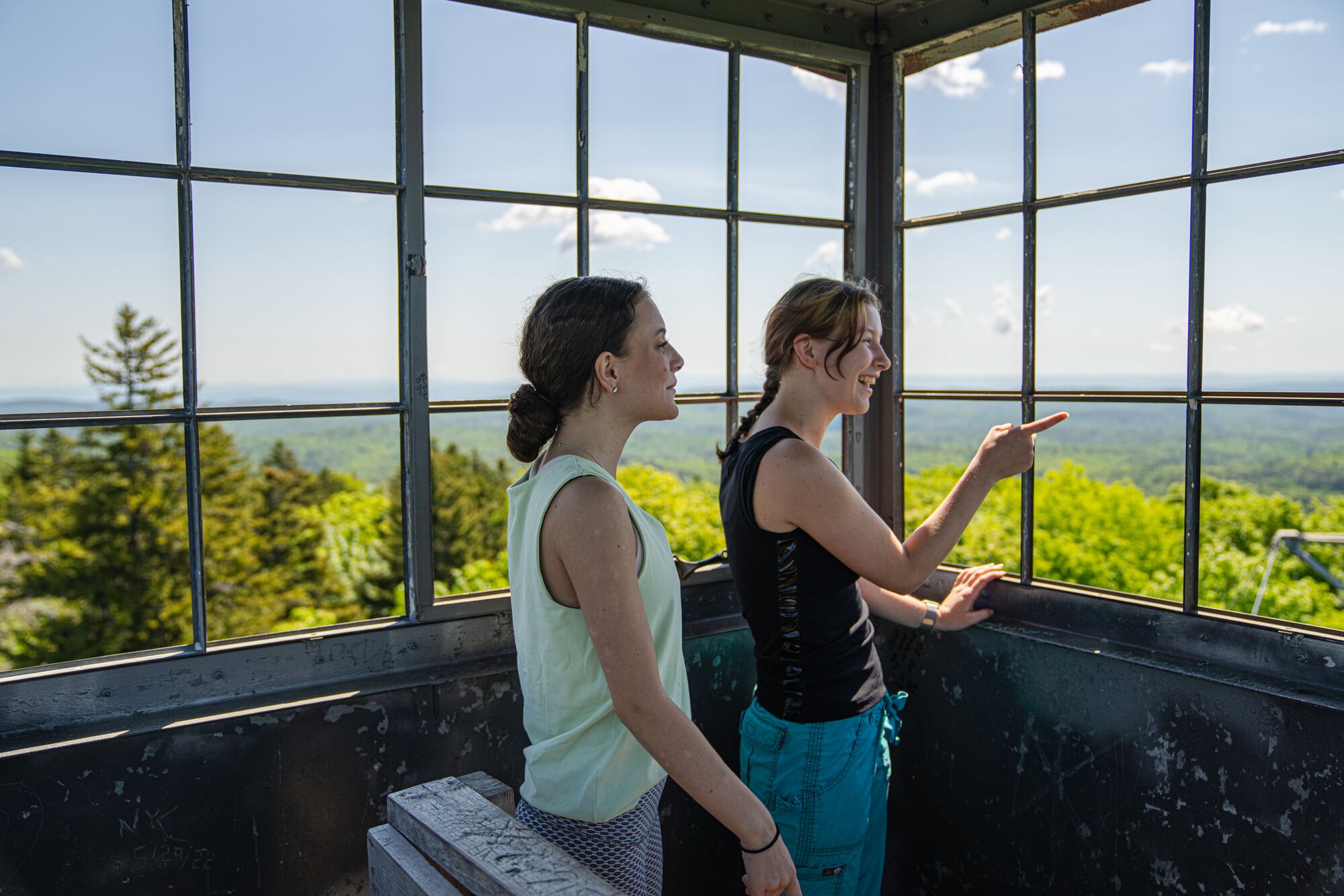 Two people at the top of a fire tower looking out at a green forest from above the treetops and pointing at the sights on a sunny summer day.