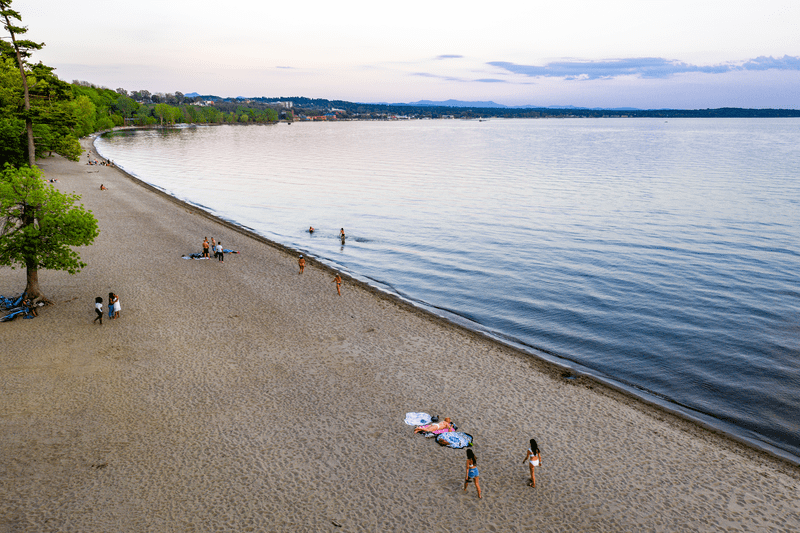 People on a sandy beach next to a lake with a city visible in the background of the image. 