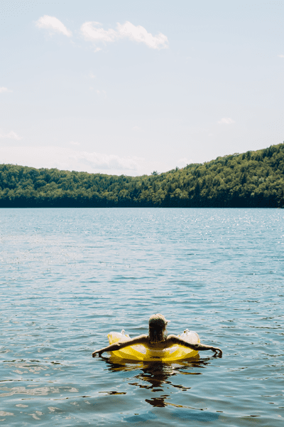 A person in a yellow floating tube on a lake in the summer. Green trees are visible surrounding.