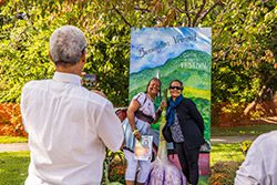 A person takes a photo of two other people in front of a sign advertising a garlic festival outside on a sunny day.