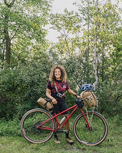 A person with a bicycle standing outside in the summer on a trail near woods.