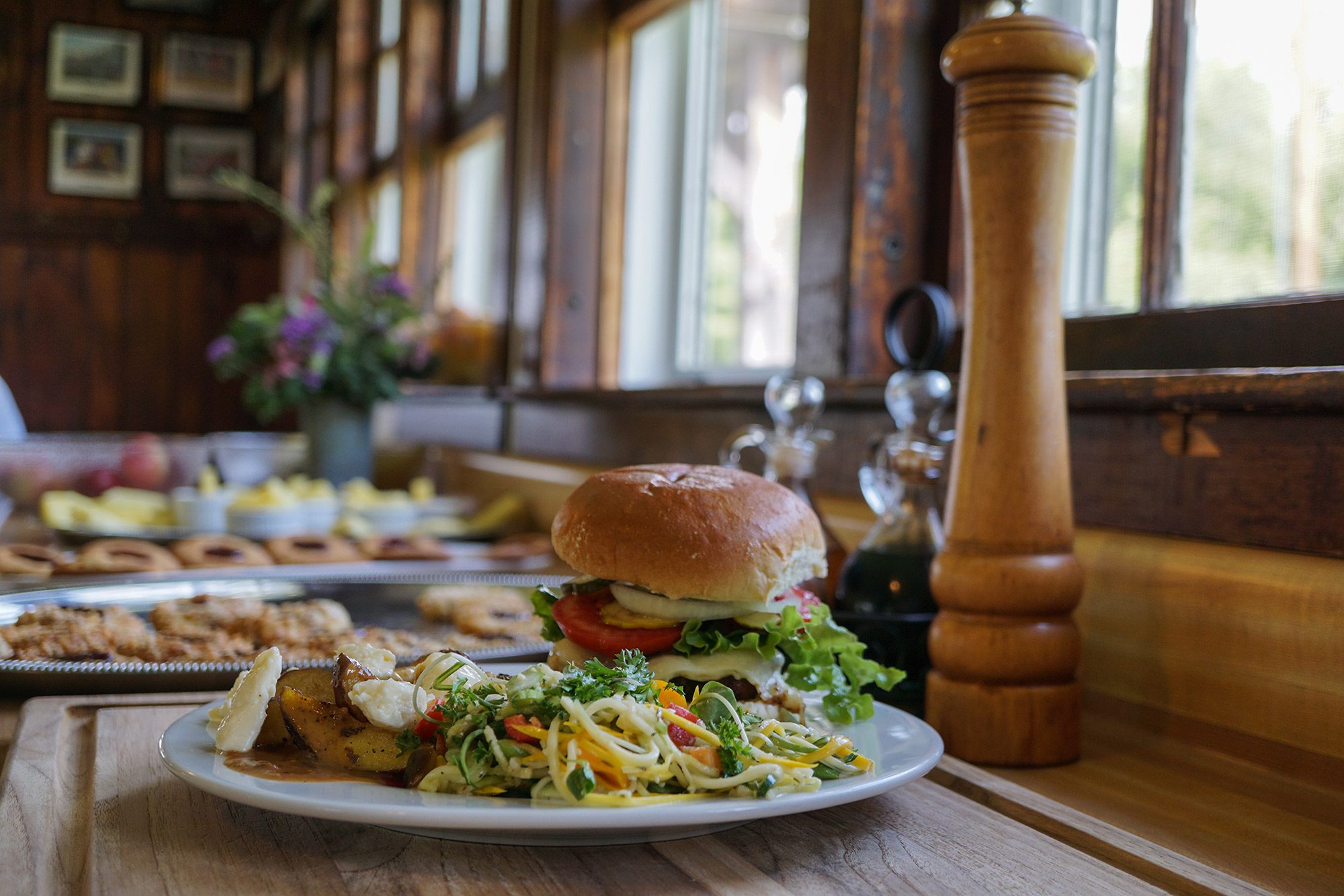 A burger with colorful toppings, greens, and potatoes on a plate by a window in the winter. 