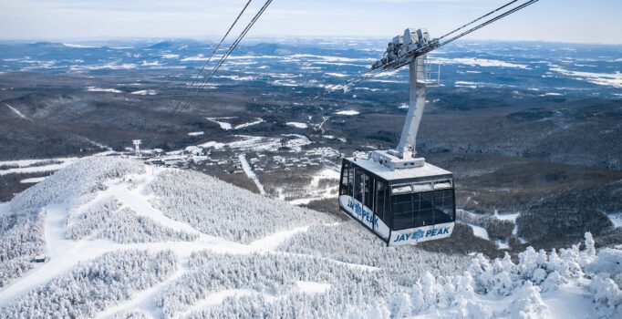 Seen from above, a ski tram that reads Jay Peak on the side, travels above snow covered ski trails.