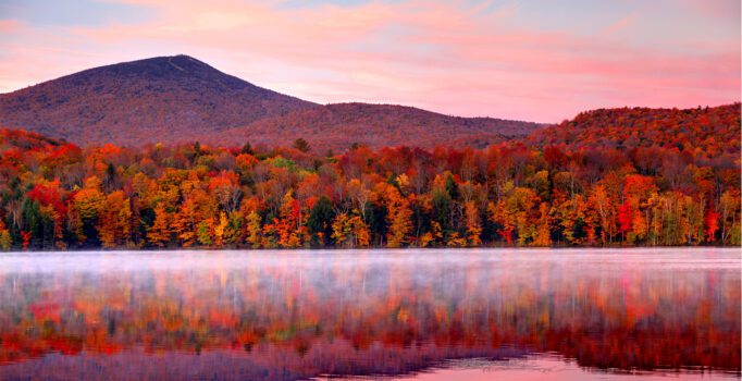 A mountain and trees in fall colors are reflecting in a calm pond, seen at sunrise.