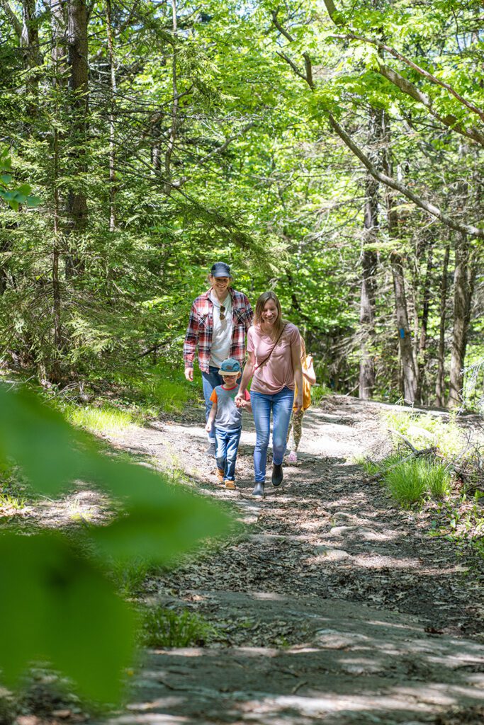 Two adults and a child hike toward the camera on an outdoor trail in the summer. 