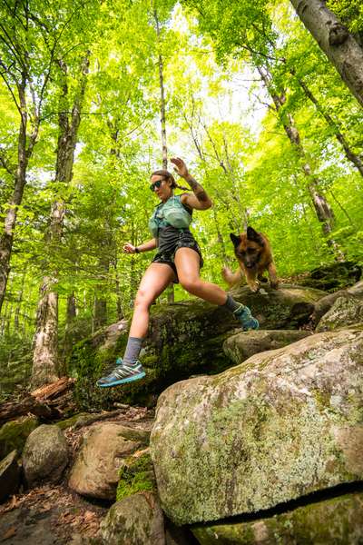 A person and a dog seen from below hiking on a rocky trail in the forest in the summer.