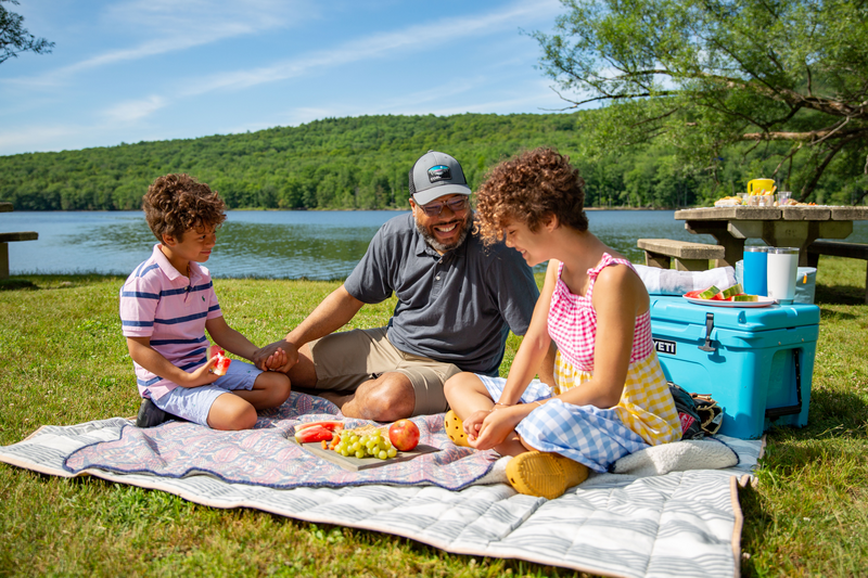 Three people sit on a blanket having a picnic next to a body of water.