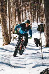 A person rides a fat bike down a snowy trail through the woods.