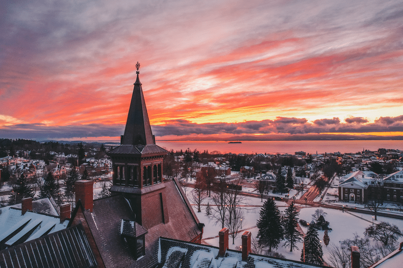 Seen from above, the sun sets in the distance across a large body of water and the buildings of a rural town in the winter.