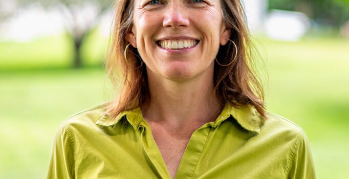 A person smiles at the camera wearing a green shirt outside on a sunny day.