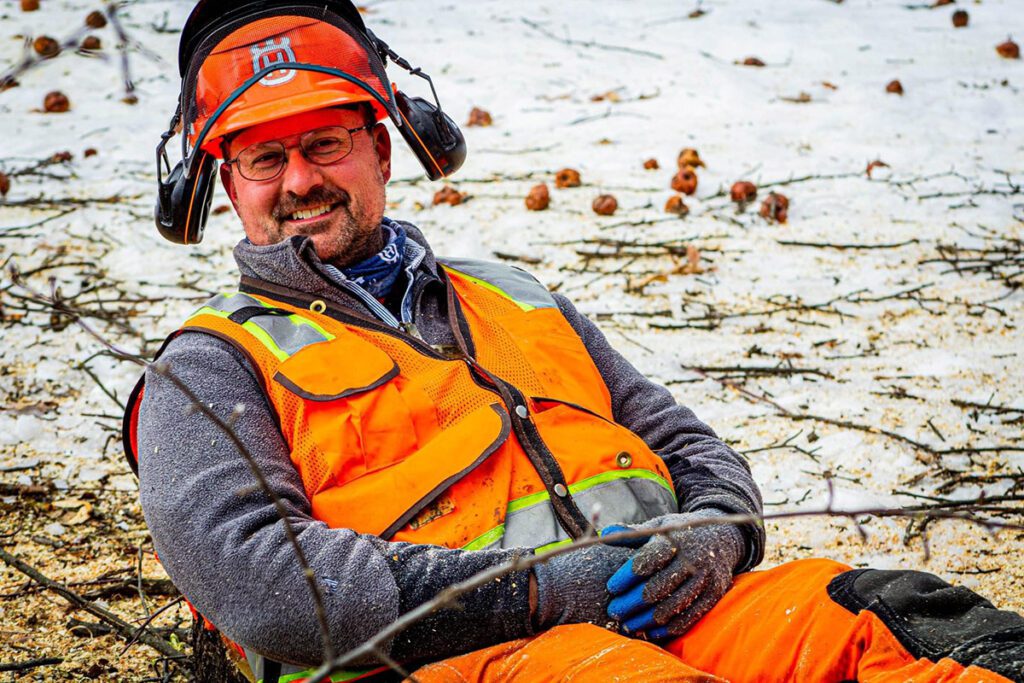 A person wearing an orange helmet, vest, and chaps sits on the ground.