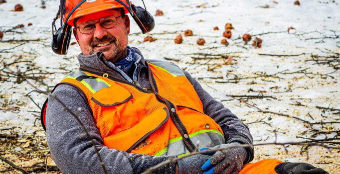 A person wearing an orange helmet, vest, and chaps sits on the ground.