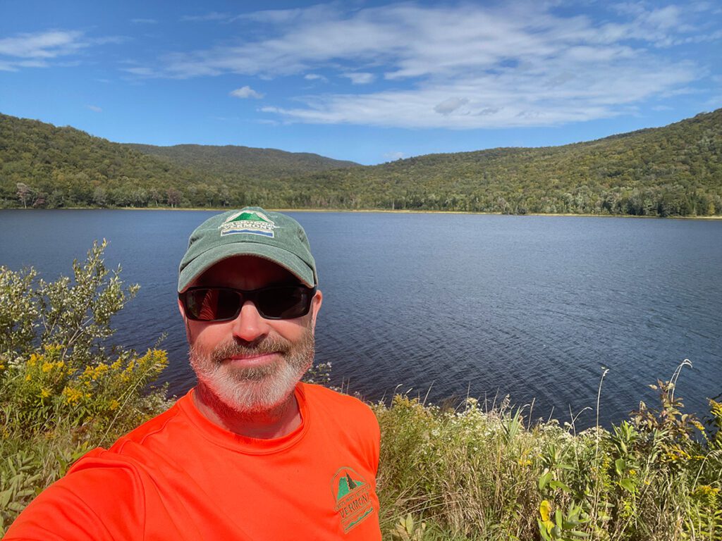 A person wearing a hat stands in front of a lake on a sunny, warm day.