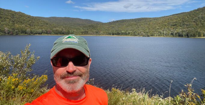 A person wearing a hat stands in front of a lake on a sunny, warm day.