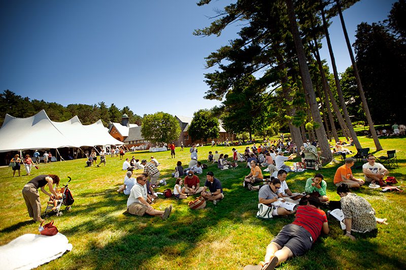 Groups of people lay on a grassy lawn with large white tents in the background.