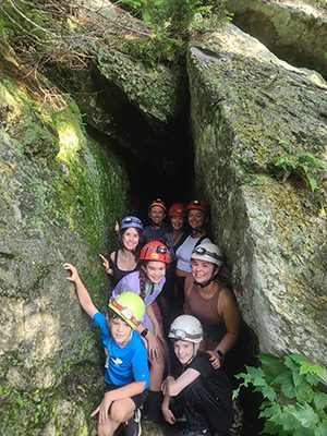 A group of people, wearing helmets and headlamps, pose for a picture just outside of a cave.