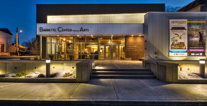 A modern building is lit up at night with performance posters on the wall and a sign above that reads Barrette Center for the Arts.