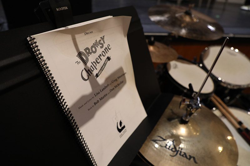 Sheet music sits on a music stand next to a drum set on a performing stage.