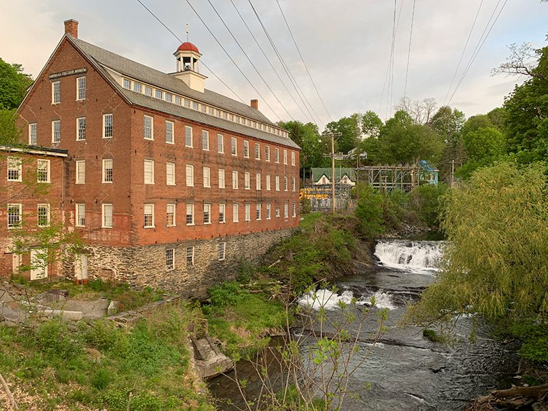 A old, brick manufacturing building overlooks the flowing water of a river.