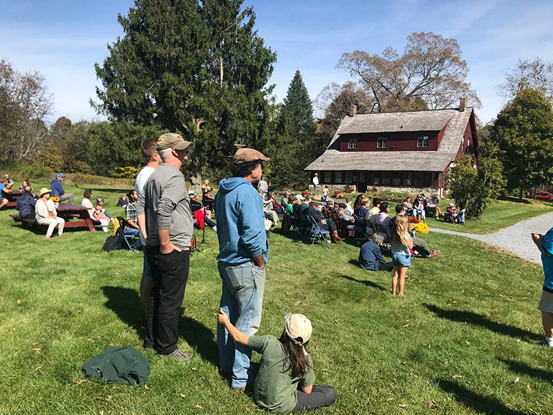 People sit outside on a grassy lawn in front of a red building to watch a presentation in the fall.