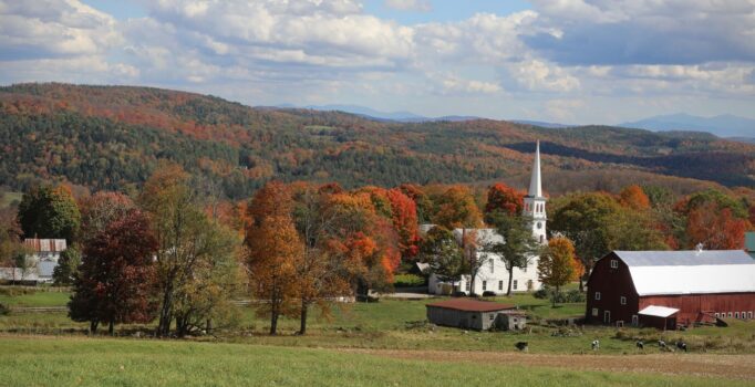 Seen from afar, a white church aside a red barn in the fall with mountains in the background.