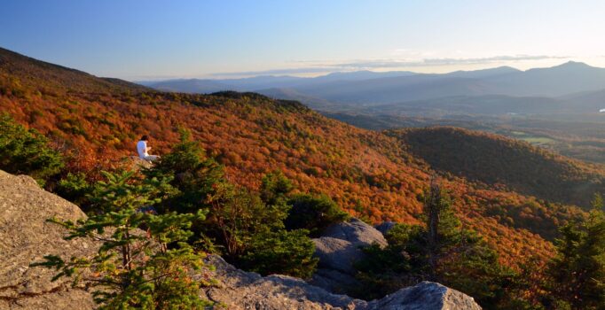 A person wearing white sits on an exposed rock atop a mountain in the fall.