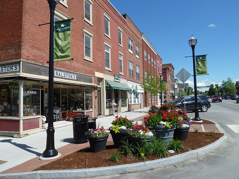 Various shops line a sidewalk, with a flower garden nestled between two light posts.