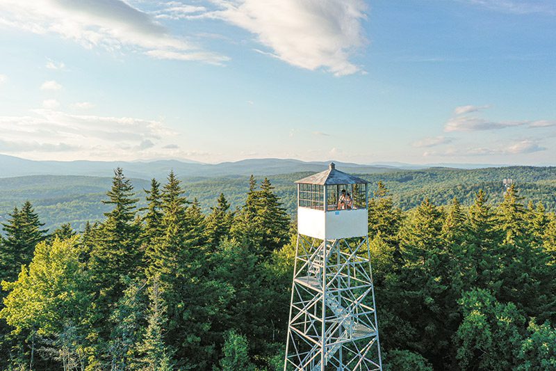 Two people stand in a fire tower that extends above the tree line at the top of a mountain on a warm day.