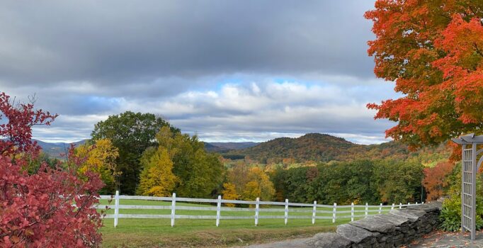 A white fence lines a green lawn and surrounded by trees with fall foliage.